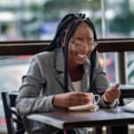 Charming African businesswoman enjoys coffee break while working at a café, embracing urban elegance.