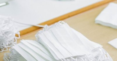 A pile of white face masks on a wooden table next to a sewing machine.